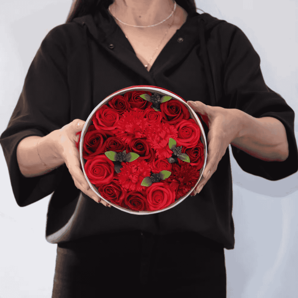 Person holding a round box filled with red soap roses against a white background