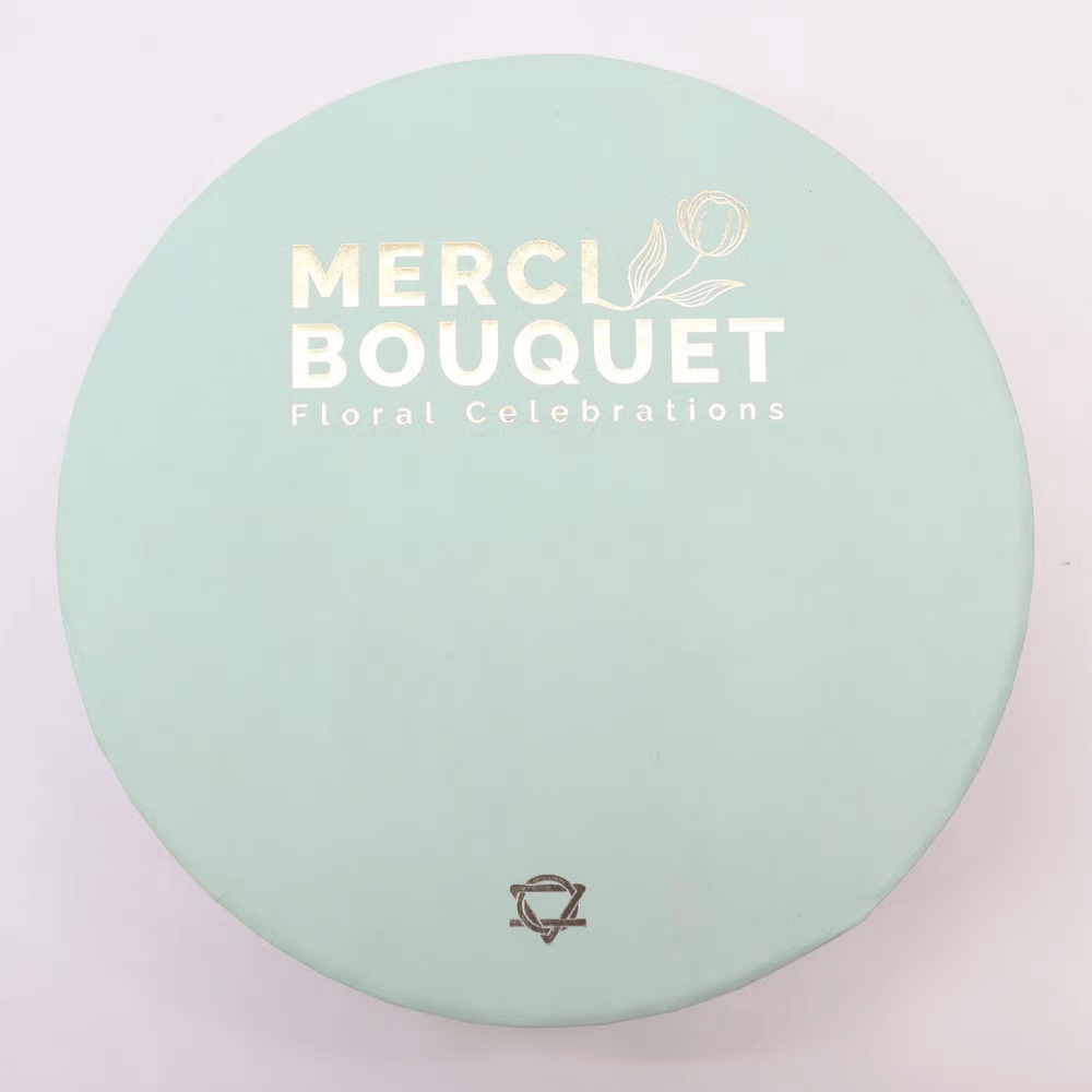 Round green box with 'MERCI BOUQUET' branding on a light pink background