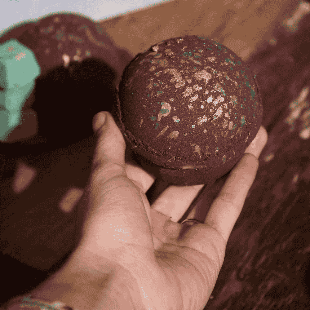 Hand holding a textured brown bath bomb with another bath bomb in the background.