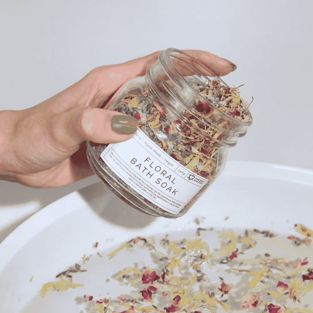 Hand holding a jar of floral bath soak with a labeled product on a white surface.