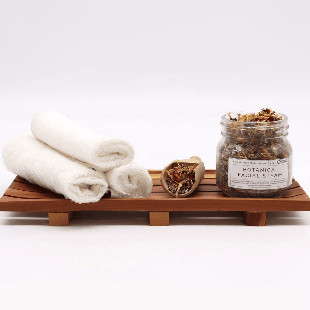 Jar of botanical facial steam with rolled towels on a wooden tray against a white background